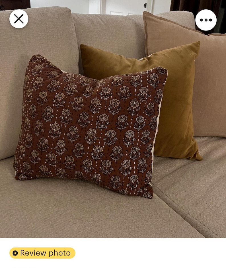 Puede incluir: Un coj&iacute;n decorativo con un estampado floral marr&oacute;n y costuras blancas. El coj&iacute;n est&aacute; sobre un sof&aacute; beige, con otros dos cojines dorados y beige. Los cojines a&ntilde;aden un toque de elegancia al sal&oacute;n.