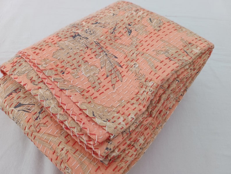 May include: Folded peach-colored quilt with a floral pattern in beige and navy blue. The quilt is hand-stitched with red and white thread, creating a textured surface. The edges are neatly finished, showcasing the craftsmanship.