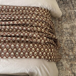 Puede incluir: Una cama con un edred&oacute;n con estampado floral marr&oacute;n y ropa de cama beige claro. El edred&oacute;n presenta un patr&oacute;n repetido de flores blancas sobre un fondo marr&oacute;n oscuro. La cama est&aacute; parcialmente cubierta con un edred&oacute;n y almohadas de color beige claro.