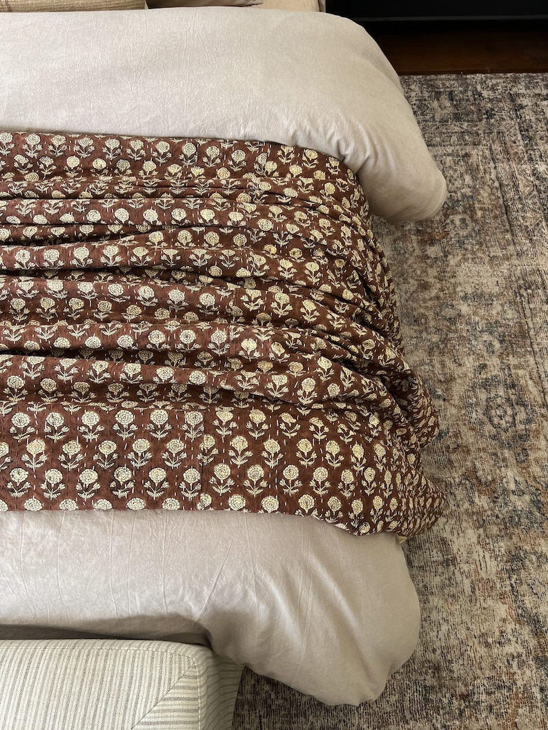 Puede incluir: Una cama con un edred&oacute;n con estampado floral marr&oacute;n y ropa de cama beige claro. El edred&oacute;n presenta un patr&oacute;n repetido de flores blancas sobre un fondo marr&oacute;n oscuro. La cama est&aacute; parcialmente cubierta con un edred&oacute;n y almohadas de color beige claro.