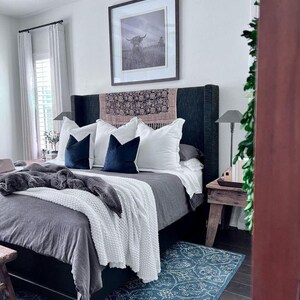 May include: A bedroom with a gray upholstered bed with white bedding and a blue throw blanket. The bed has a black headboard with a patterned throw pillow. A blue rug with a white pattern is on the floor. A wooden side table with a lamp is next to the bed.