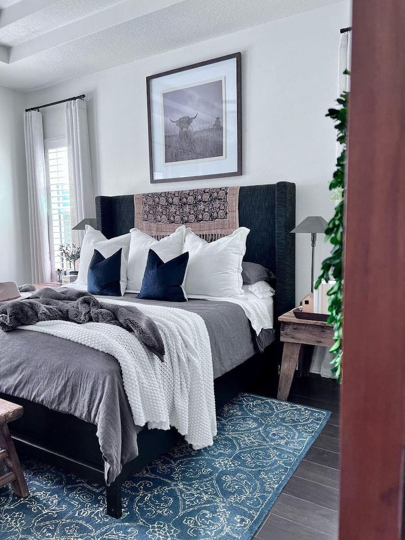 May include: A bedroom with a gray upholstered bed with white bedding and a blue throw blanket. The bed has a black headboard with a patterned throw pillow. A blue rug with a white pattern is on the floor. A wooden side table with a lamp is next to the bed.