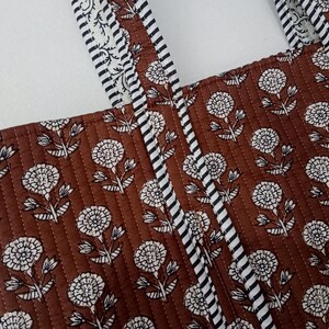 Puede incluir: Primer plano de un bolso tote acolchado marr&oacute;n con un estampado floral blanco. El bolso tiene dos correas con un dise&ntilde;o a rayas blancas y negras. El estampado floral se repite en la superficie del bolso.