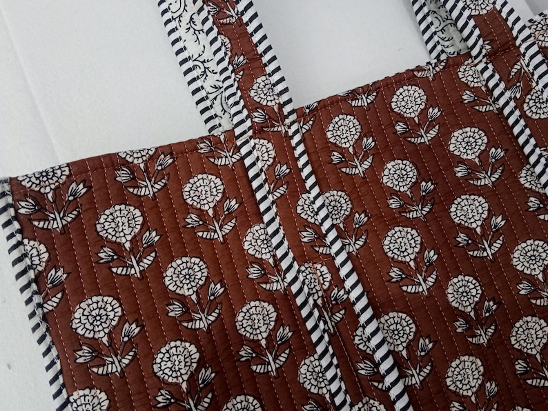 Puede incluir: Primer plano de un bolso tote acolchado marr&oacute;n con un estampado floral blanco. El bolso tiene dos correas con un dise&ntilde;o a rayas blancas y negras. El estampado floral se repite en la superficie del bolso.