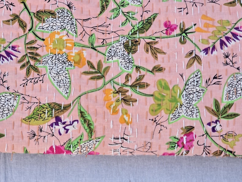 May include: A pink floral fabric with a repeating pattern of leaves and flowers in shades of green, yellow, purple, and pink. The design is stitched with white thread, creating a quilted effect. The bottom edge is a solid gray.