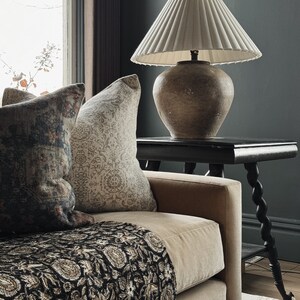 May include: A beige sofa with a black and cream patterned throw blanket. Two pillows are on the sofa, one with a floral pattern and the other with a paisley pattern. A table with a lamp sits next to the sofa. The lamp has a white pleated shade and a brown ceramic base.