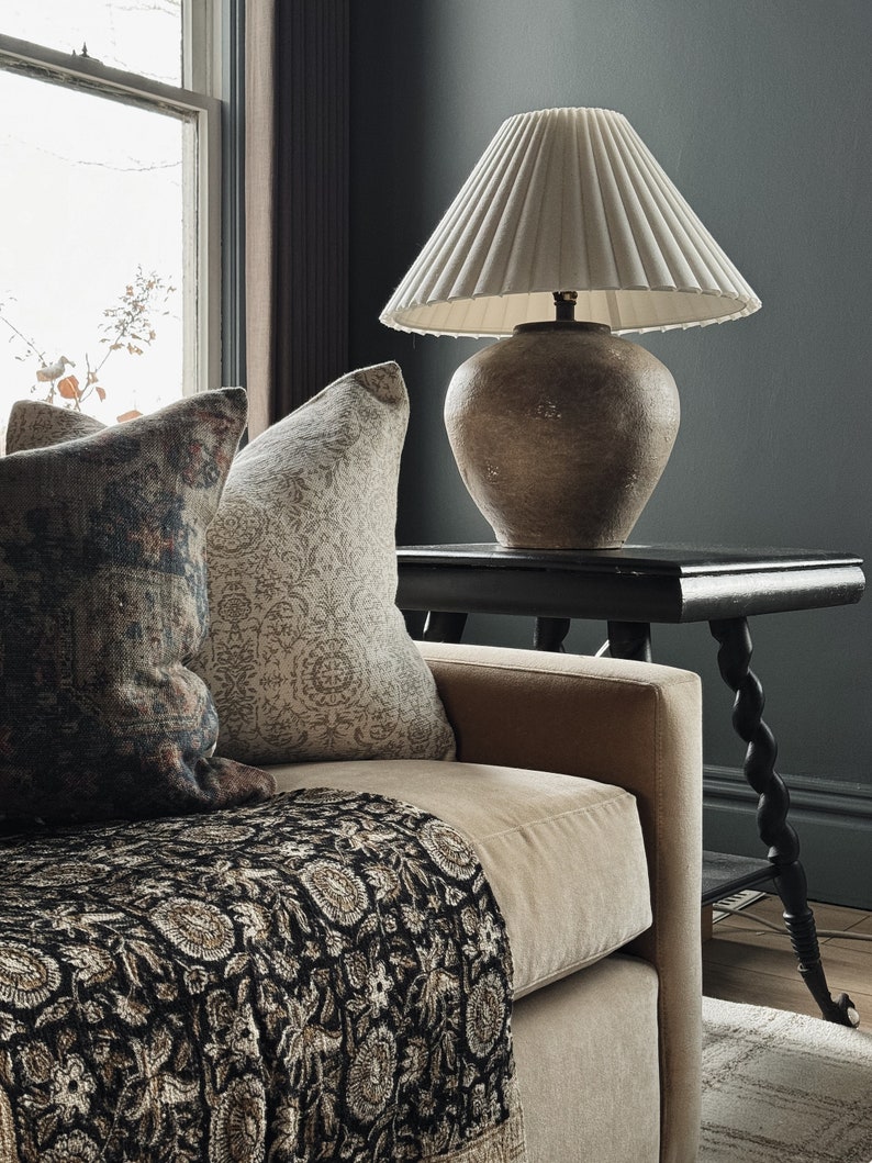May include: A beige sofa with a black and cream patterned throw blanket. Two pillows are on the sofa, one with a floral pattern and the other with a paisley pattern. A table with a lamp sits next to the sofa. The lamp has a white pleated shade and a brown ceramic base.