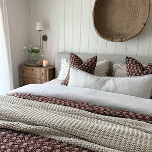 Puede incluir: Un dormitorio con una cama con almohadas blancas y burdeos con estampado floral y un edred&oacute;n a juego. Una cesta tejida cuelga de la pared sobre la cama. Una mesita de noche de madera con una vela y flores est&aacute; a la izquierda.