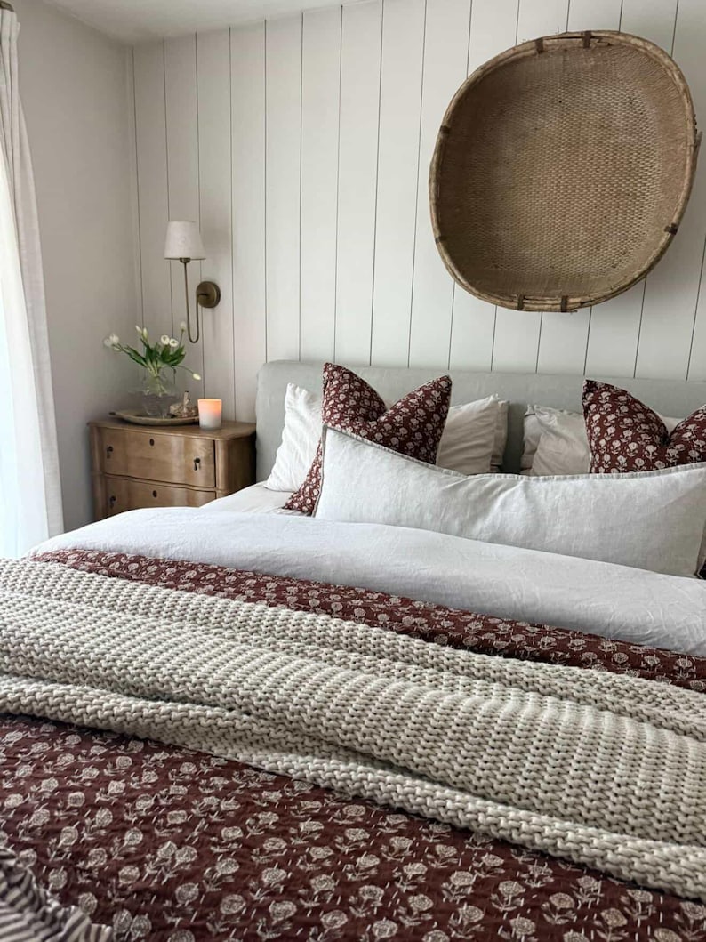 Puede incluir: Un dormitorio con una cama con almohadas blancas y burdeos con estampado floral y un edred&oacute;n a juego. Una cesta tejida cuelga de la pared sobre la cama. Una mesita de noche de madera con una vela y flores est&aacute; a la izquierda.