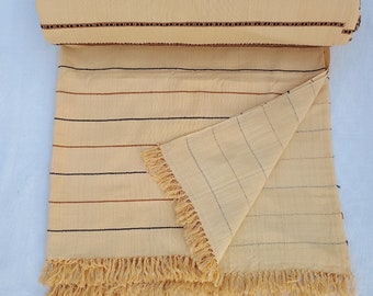 Puede incluir: Textil beige con detalles de rayas marrones y negras y un borde con flecos. La parte superior está enrollada, revelando un borde detallado. La tela tiene un aspecto suave y texturizado, adecuada para una manta o un cubrecama.
