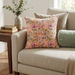 May include: A square, floral-patterned pillow in shades of pink, green, and purple rests on a beige sofa. The pillow features a detailed botanical design. A green bolster pillow and a beige throw blanket are also visible.