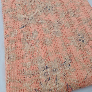 May include: Peach-colored, floral-patterned quilt with intricate stitching. The quilt features a repeating design of stylized flowers and leaves in shades of beige and navy blue. The stitching is in a contrasting red thread, creating a textured effect.