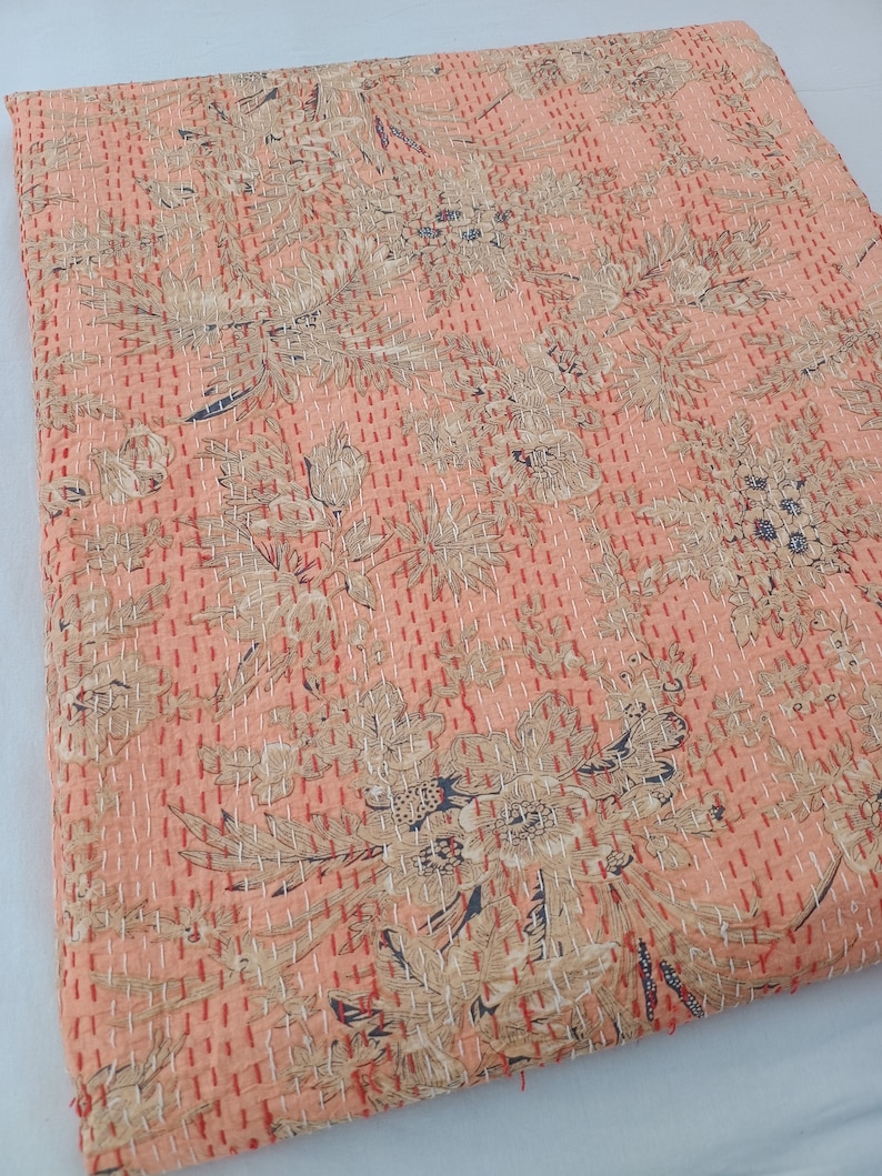 May include: Peach-colored, floral-patterned quilt with intricate stitching. The quilt features a repeating design of stylized flowers and leaves in shades of beige and navy blue. The stitching is in a contrasting red thread, creating a textured effect.