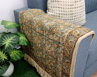 May include: A blue sofa with a floral patterned throw and a decorative cushion. The throw has a green background with yellow flowers and a fringed edge. The cushion has a white and grey dotted pattern.