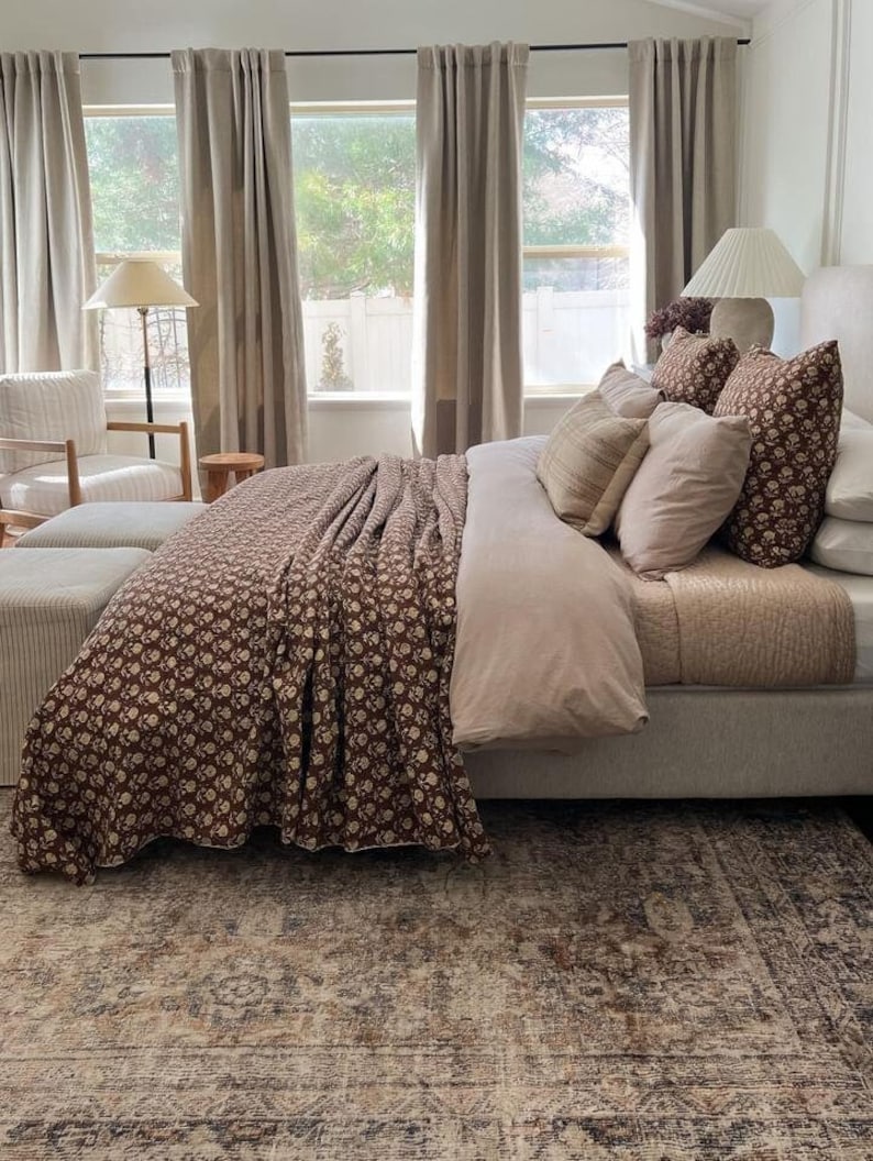 Puede incluir: Interior de dormitorio con una cama vestida con un edred&oacute;n floral marr&oacute;n y cojines. La cama tiene una funda n&oacute;rdica rosa claro y varias almohadas decorativas. Una alfombra estampada y cortinas neutras crean un ambiente c&aacute;lido y acogedor.