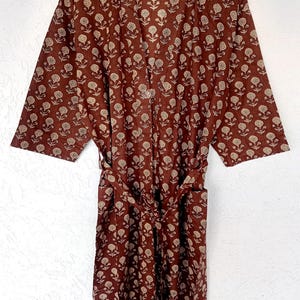 Puede incluir: Una bata marr&oacute;n con un estampado floral, colgada de una percha de madera. La bata tiene un cintur&oacute;n y un dise&ntilde;o repetido de flores blancas. La tela parece ligera, adecuada para descansar o como abrigo.