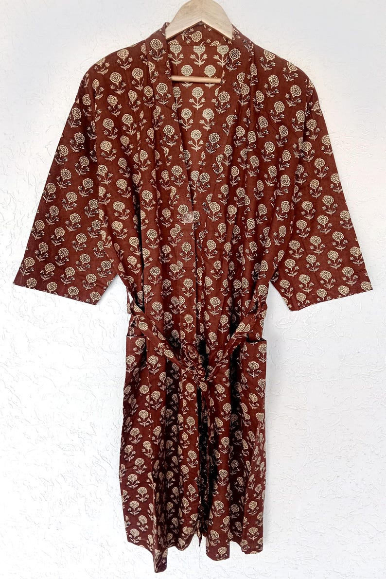 Puede incluir: Una bata marr&oacute;n con un estampado floral, colgada de una percha de madera. La bata tiene un cintur&oacute;n y un dise&ntilde;o repetido de flores blancas. La tela parece ligera, adecuada para descansar o como abrigo.