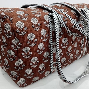Puede incluir: Un bolso tote acolchado marr&oacute;n con un estampado floral blanco. El bolso tiene dos asas con un dise&ntilde;o de rayas blancas y negras. Los bordes del bolso est&aacute;n adornados con un estampado floral blanco.