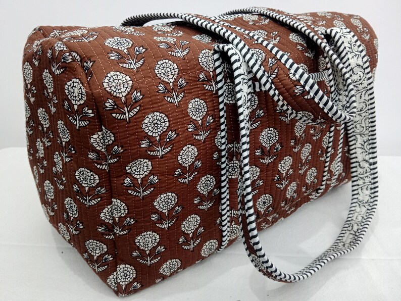 Puede incluir: Un bolso tote acolchado marr&oacute;n con un estampado floral blanco. El bolso tiene dos asas con un dise&ntilde;o de rayas blancas y negras. Los bordes del bolso est&aacute;n adornados con un estampado floral blanco.