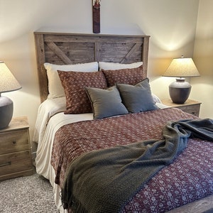 May include: A rustic wooden bed frame with a brown and white patterned quilt and a green throw blanket. The bed has a wooden headboard with a cross above it. There are two bedside tables with lamps on them.