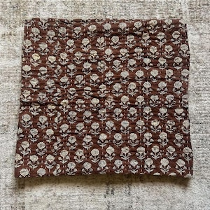 Puede incluir: Un edred&oacute;n marr&oacute;n con estampado floral. El edred&oacute;n presenta un patr&oacute;n repetitivo de flores blancas con costuras detalladas. La tela tiene una textura acolchada y el dise&ntilde;o general es sim&eacute;trico. El edred&oacute;n es rectangular.
