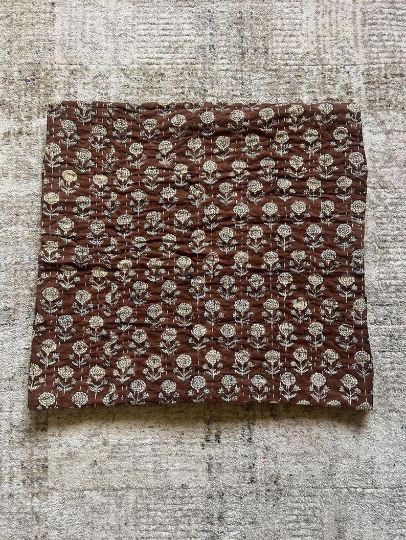 Puede incluir: Un edred&oacute;n marr&oacute;n con estampado floral. El edred&oacute;n presenta un patr&oacute;n repetitivo de flores blancas con costuras detalladas. La tela tiene una textura acolchada y el dise&ntilde;o general es sim&eacute;trico. El edred&oacute;n es rectangular.