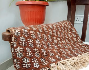Puede incluir: Una manta de tiro marrón con un patrón floral blanco y ribete de flecos. La manta está drapeada sobre una silla de madera.
