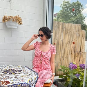 May include: A red and white gingham print two-piece set consisting of a short-sleeved top with a front tie closure and matching pants. The top features a scoop neckline and the fabric has a small checkered pattern throughout the entire garment.