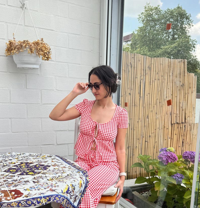 May include: A red and white gingham print two-piece set consisting of a short-sleeved top with a front tie closure and matching pants. The top features a scoop neckline and the fabric has a small checkered pattern throughout the entire garment.