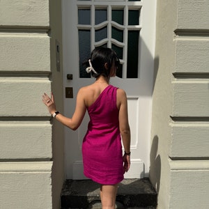 May include: A woman wearing a one-shoulder fuchsia dress stands in front of a white door with a decorative window. She is wearing a white hair clip and a watch on her wrist.