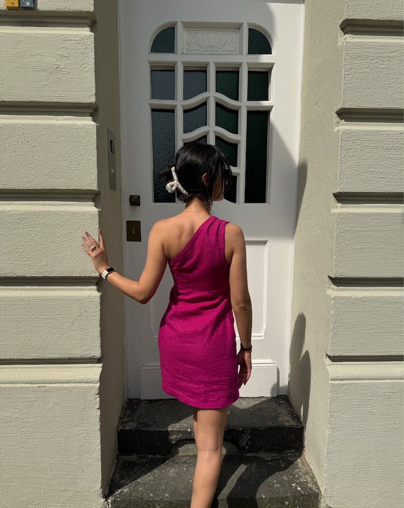 May include: A woman wearing a one-shoulder fuchsia dress stands in front of a white door with a decorative window. She is wearing a white hair clip and a watch on her wrist.