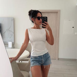 May include: A white sleeveless top with a square neckline and fitted waist is paired with denim shorts. The shorts are light blue with a frayed hem. The person is wearing black sunglasses and holding a black phone.