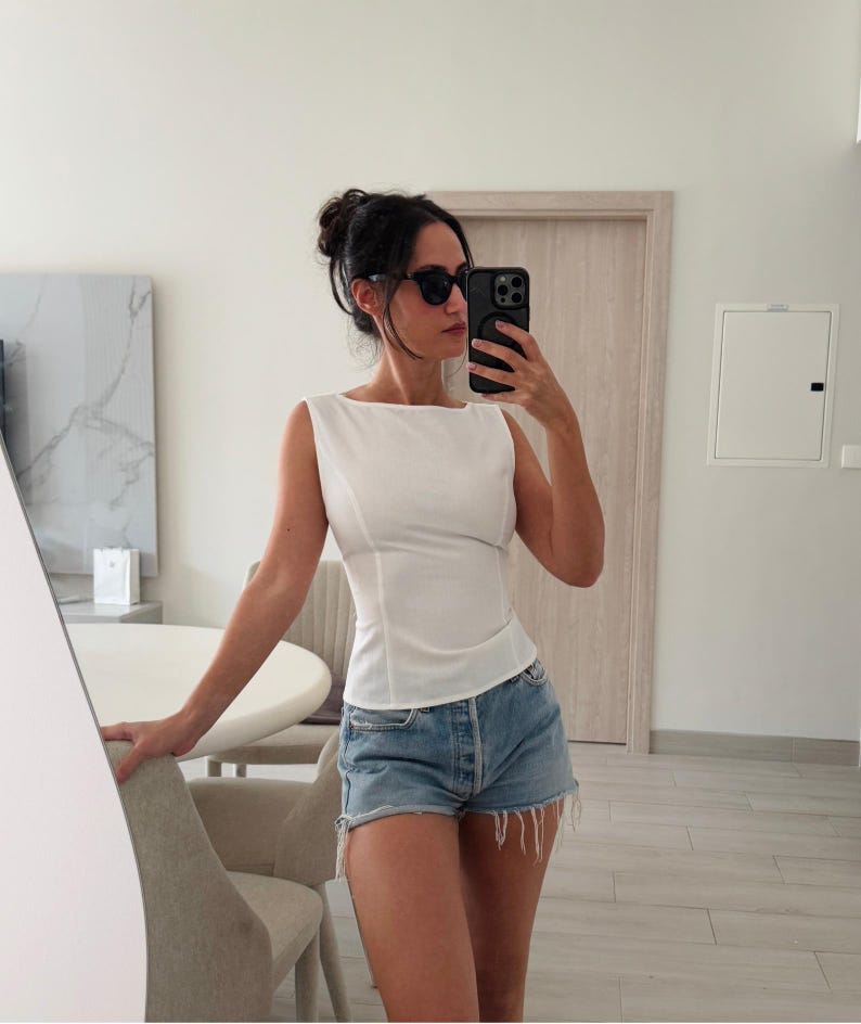 May include: A white sleeveless top with a square neckline and fitted waist is paired with denim shorts. The shorts are light blue with a frayed hem. The person is wearing black sunglasses and holding a black phone.