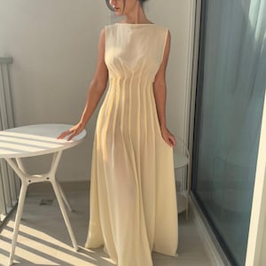 May include: A sleeveless, floor-length, pale yellow dress. The dress features vertical pleats on the bodice and a flowing skirt. The dress has a boat neckline and is made of a lightweight, semi-sheer fabric. The dress is worn outdoors on a sunny day.