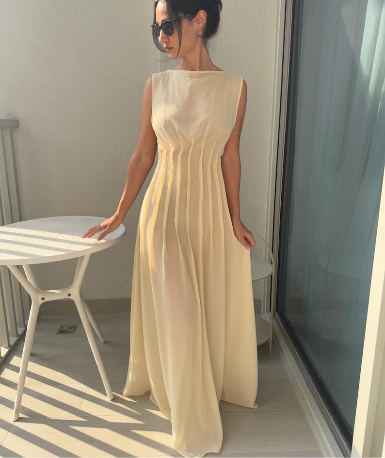 May include: A sleeveless, floor-length, pale yellow dress. The dress features vertical pleats on the bodice and a flowing skirt. The dress has a boat neckline and is made of a lightweight, semi-sheer fabric. The dress is worn outdoors on a sunny day.