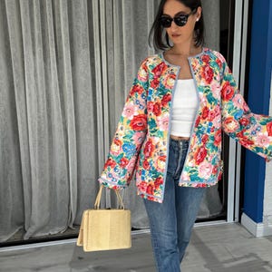 May include: A floral print jacket with red, pink, blue, and yellow flowers. The jacket has a light blue trim and is worn over a white top and blue jeans. A light yellow handbag is held in the hand.