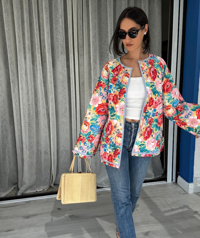 May include: A floral print jacket with red, pink, blue, and yellow flowers. The jacket has a light blue trim and is worn over a white top and blue jeans. A light yellow handbag is held in the hand.