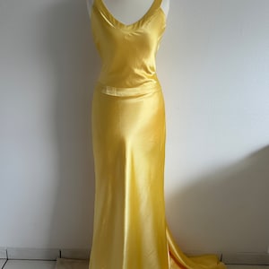 Puede incluir: Un vestido largo de sat&eacute;n amarillo con escote en V y un corpi&ntilde;o ajustado. El vestido tiene una falda fluida y una cola.