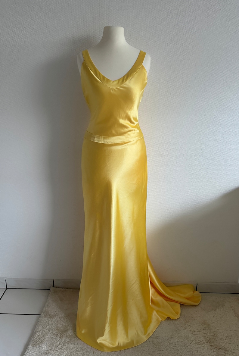 Puede incluir: Un vestido largo de sat&eacute;n amarillo con escote en V y un corpi&ntilde;o ajustado. El vestido tiene una falda fluida y una cola.