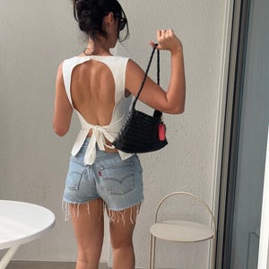 May include: A white sleeveless top with a large back cutout and a tie closure, paired with light-wash denim shorts. A black woven handbag with a red tag is held in the hand. The outfit is completed with sunglasses and a hair bun.