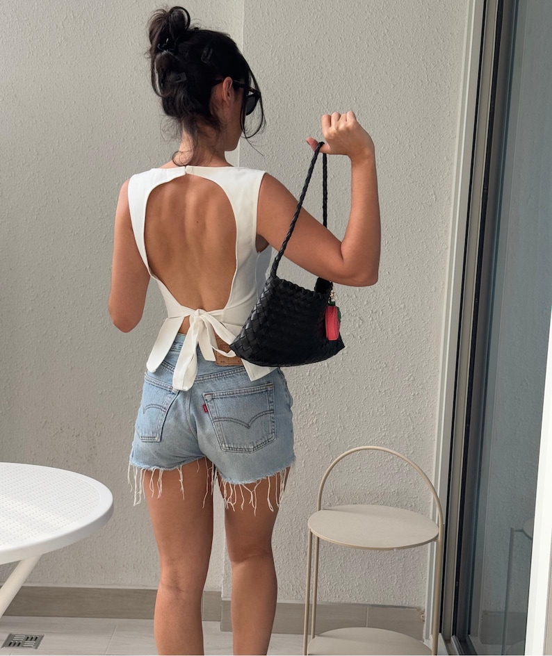 May include: A white sleeveless top with a large back cutout and a tie closure, paired with light-wash denim shorts. A black woven handbag with a red tag is held in the hand. The outfit is completed with sunglasses and a hair bun.