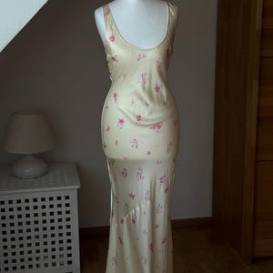 May include: A long, sleeveless, cream-colored satin dress with a floral print in shades of pink and green. The dress has a scoop neck and a fitted bodice. The skirt is flowing and has a slight train.