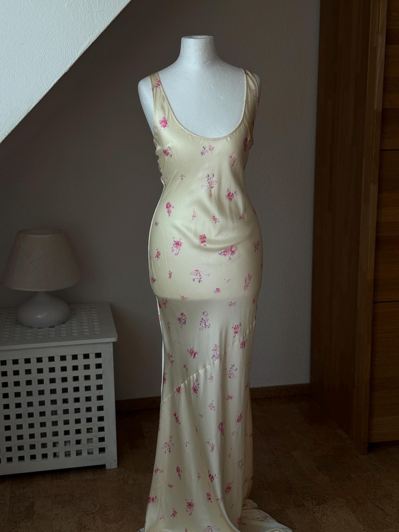 May include: A long, sleeveless, cream-colored satin dress with a floral print in shades of pink and green. The dress has a scoop neck and a fitted bodice. The skirt is flowing and has a slight train.
