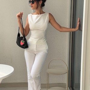 May include: A woman in a white sleeveless top and matching pants, accessorized with black sunglasses and a black handbag with a strawberry charm. The outfit is paired with nude sandals. A small, two-tiered side table is visible.
