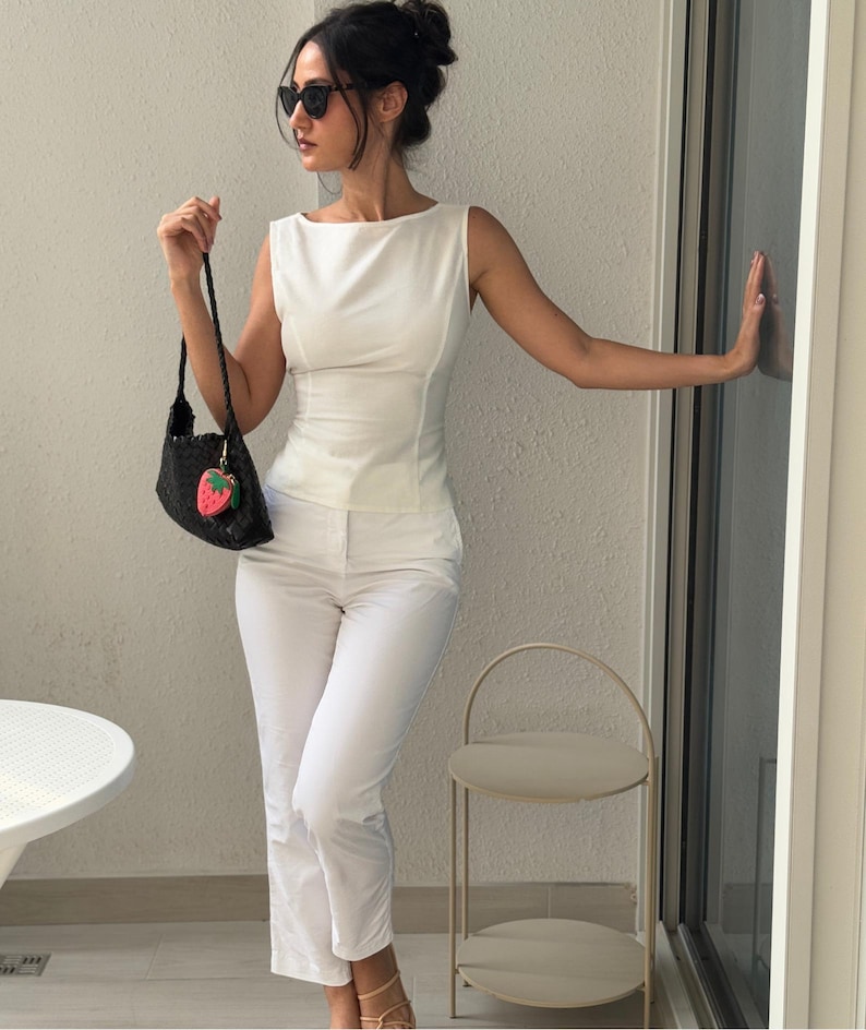 May include: A woman in a white sleeveless top and matching pants, accessorized with black sunglasses and a black handbag with a strawberry charm. The outfit is paired with nude sandals. A small, two-tiered side table is visible.
