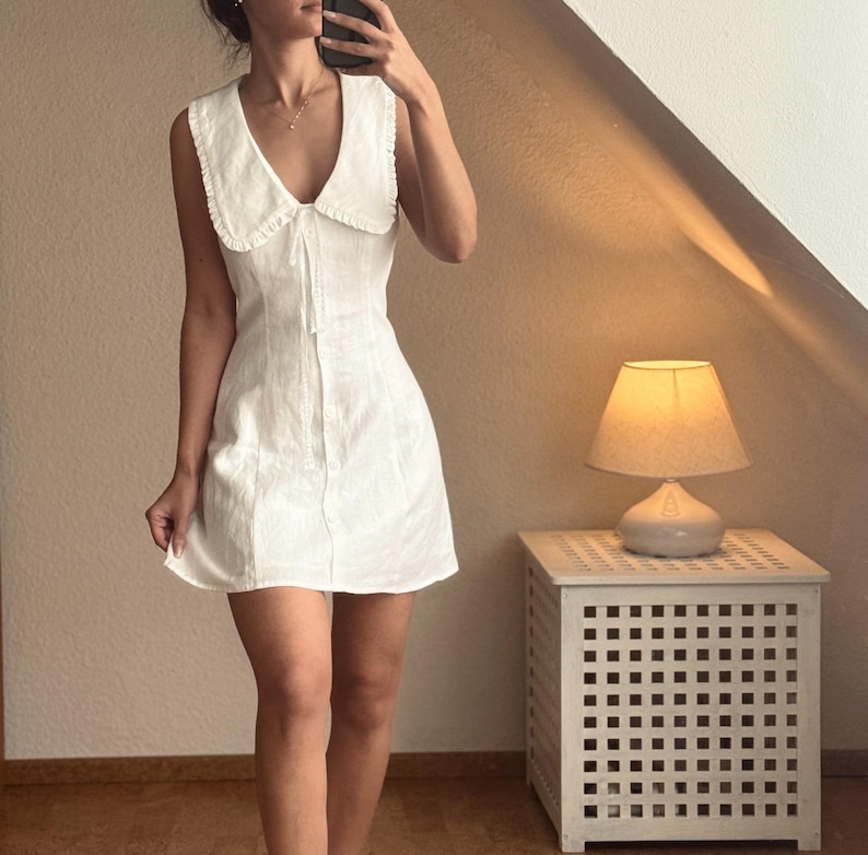 May include: A sleeveless white mini dress featuring a large collar with ruffled edges, a deep V-neckline, and a front button closure. The dress has a fitted bodice that flares slightly at the hem, crafted from a textured fabric with a clean finish.