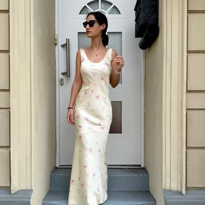 May include: A woman wearing a white maxi dress with a pink floral pattern. She is standing on a set of stairs in front of a white door.