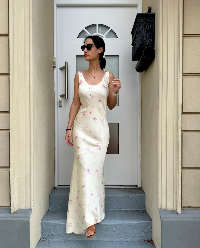 May include: A woman wearing a white maxi dress with a pink floral pattern. She is standing on a set of stairs in front of a white door.