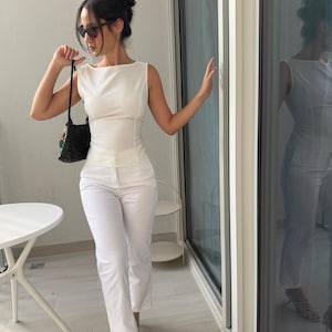 May include: A sleeveless white top and matching white pants. The top has a boat neck and fitted waist. A black woven handbag with colorful accents is held in the left hand. The person is wearing sunglasses and standing on a balcony.
