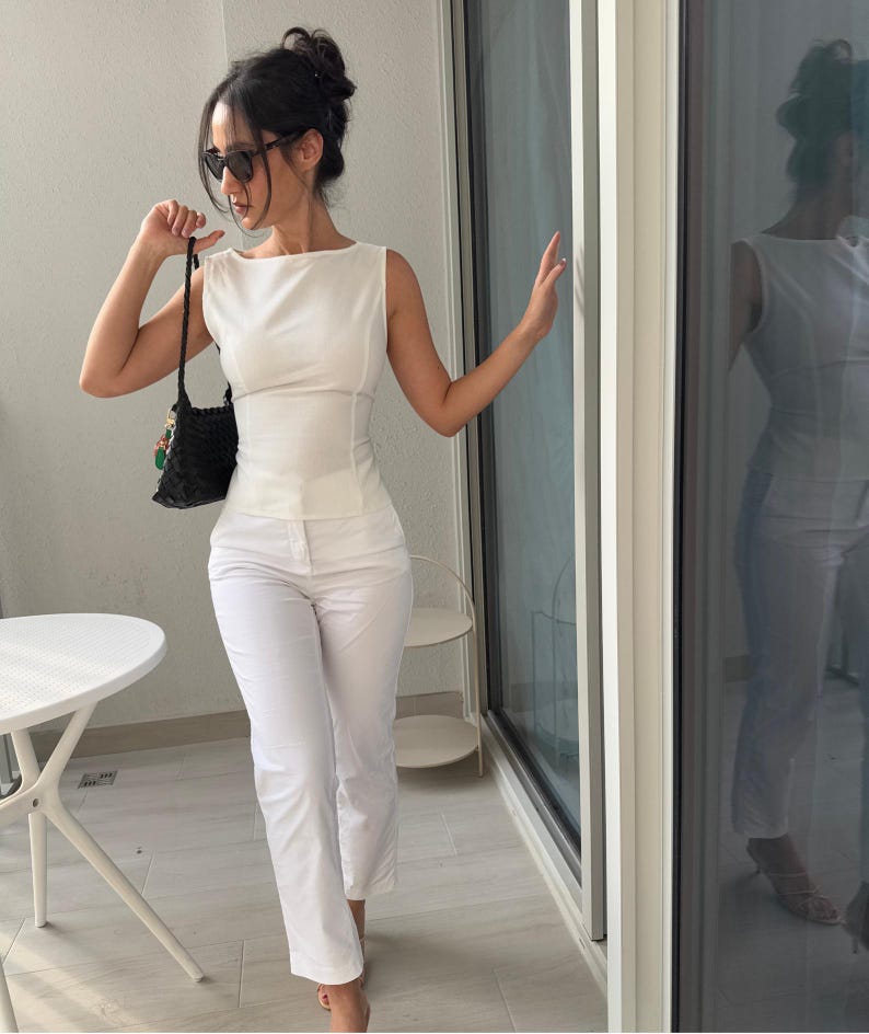 May include: A sleeveless white top and matching white pants. The top has a boat neck and fitted waist. A black woven handbag with colorful accents is held in the left hand. The person is wearing sunglasses and standing on a balcony.